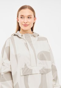 Krämfärgad regnjacka med abstrakta grå mönster, huva och en framficka. Tillverkad av lätt, vattenavvisande material.