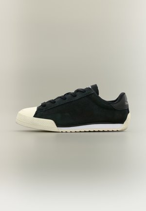 Zapatilla negra de corte bajo con puntera de caucho blanca, cordones negros y suela blanca sobre un fondo neutro, vista lateral.