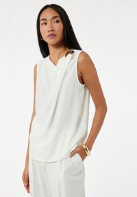 Biała bluzka bez rękawów o drapowanej konstrukcji, z złotym okrągłym akcentem na ramieniu. Gładka tekstura, w zestawieniu z luźnymi białymi spodniami.