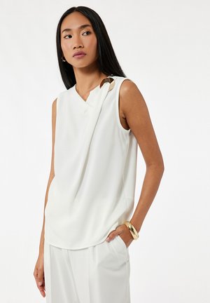 Biała bluzka bez rękawów o drapowanej konstrukcji, z złotym okrągłym akcentem na ramieniu. Gładka tekstura, w zestawieniu z luźnymi białymi spodniami.