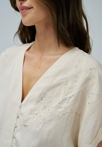 Blusa beige chiaro con scollo a V, chiusura con bottoni e preziosa ricamatura floreale con perline e paillettes lungo lo scollo e le spalle.