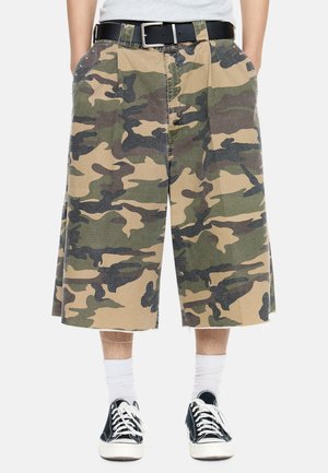 Camo-mønstrede shorts med vide ben og frynset kant, båret med sort bælte, hvide sokker og sorte lave sneakers.