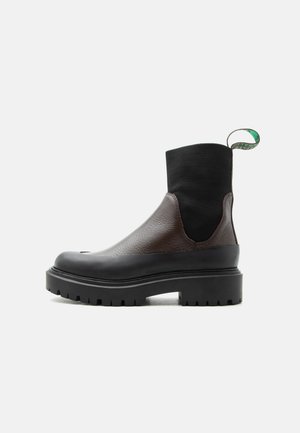 Čierna a hnedá členková kožená topánka s elastickým textilným zvrškom, robustnou podrážkou s hrubým vzorom a zeleným sťahovacím remienkom na bielom pozadí.