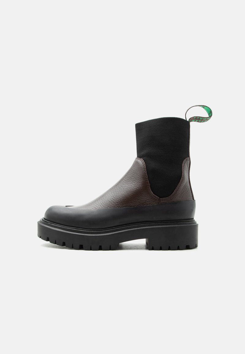 Μαύρο και καφέ δερμάτινο μποτάκι αστραγάλου με ελαστικό επάνω μέρος από ύφασμα, χοντρή σόλα με αυλακώσεις και πράσινη θηλιά τράβηγμα, σε λευκό φόντο.