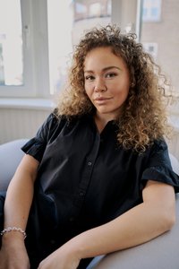 Jeune femme aux cheveux bouclés portant une chemise noire, assise dans une pièce lumineuse près d'une fenêtre, regardant calmement la caméra.