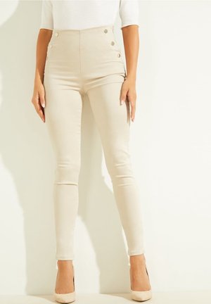 Mujer con pantalones pitillo beige de cintura alta con botones decorativos, combinados con tacones altos beige, de pie frente a un fondo claro.