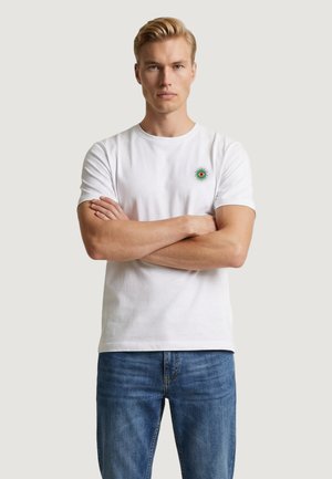 Giovane con le braccia incrociate che indossa una t-shirt bianca con un piccolo emblema verde e rosso sul petto e jeans blu, sfondo neutro.