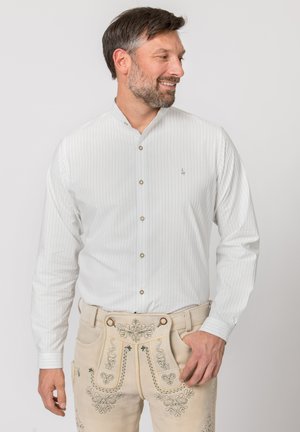 Mann trägt ein weißes Nadelstreifenhemd und beige traditionelle bestickte Lederhosen, schaut nach rechts und lächelt.