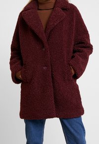 Cappotto in stile montone color bordeaux con colletto a berretto e chiusura frontale con bottoni. Presenta tasche laterali e una vestibilità comoda.