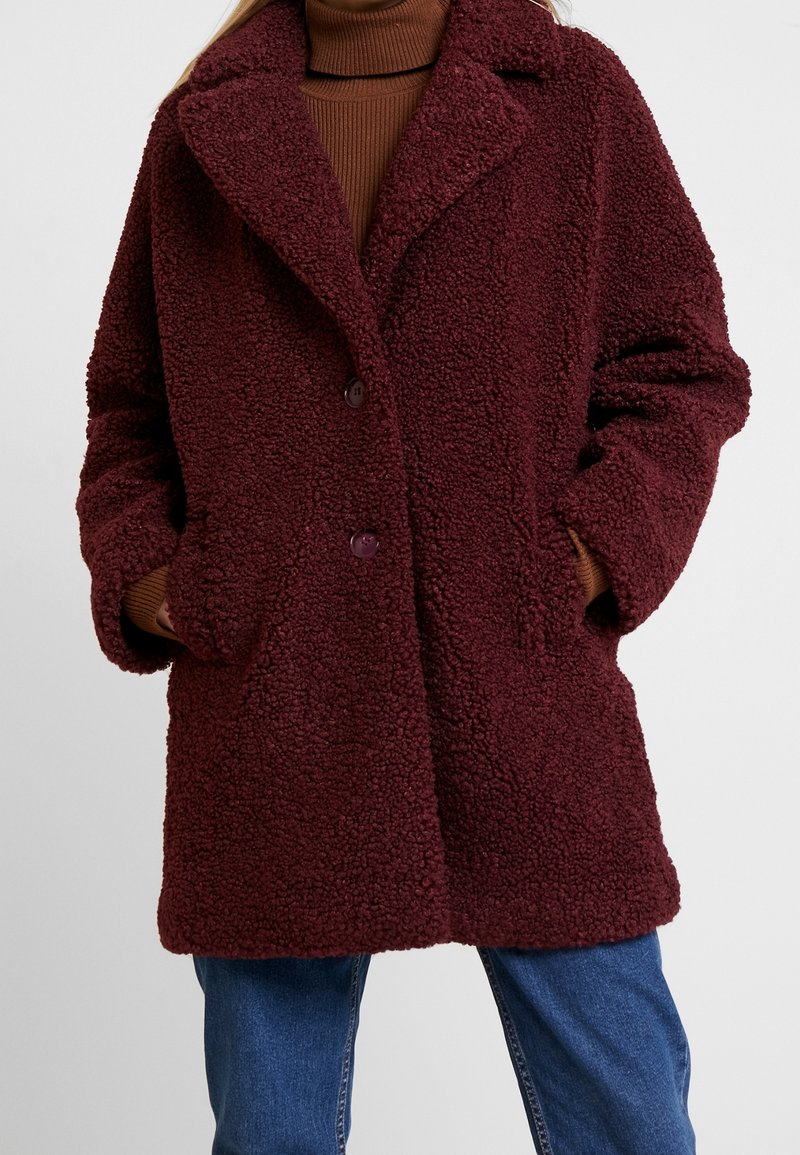 Cappotto in stile montone color bordeaux con colletto a berretto e chiusura frontale con bottoni. Presenta tasche laterali e una vestibilità comoda.