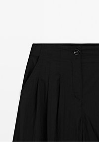 Pantalon noir taille haute avec plis devant, poches latérales et une fermeture par un bouton noir unique à la taille.