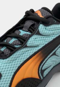 Tyrkysově modrá a černá sportovní obuv s texturovaným síťovaným svrškem, oranžovým akcentem a černými tkaničkami. Výrazné logo v blízkosti paty.