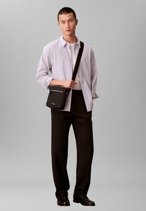 Bolso bandolera negro con tela texturizada, con un bolsillo con cremallera. El modelo lleva una camisa blanca de rayas, pantalones negros y zapatos pulidos.