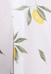 Witte stof met een patroon van groene bladeren en gele citroenen, met twee witte knopen op de voorkant van een shirt.