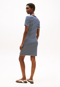 Woman wearing a navy and white striped short-sleeve dress and white sandals, standing and looking over her shoulder against a white background.