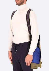 Dolcevita crema, bretelle nere con scritte blu, pantaloni navy con coulisse, tiene una borsa verde con interno blu. Design minimalista.