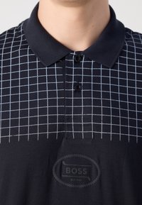 Gros plan d'une personne portant un polo noir avec un motif quadrillé blanc et un écusson avec le logo Boss sur la poitrine.