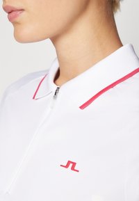 Bjela polo majica s ovratnikom na zatvarač, s crvenim detaljima i malim crvenim logotipom. Tkanina ima glatku teksturu i dobro se prilagođava tijelu.