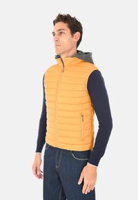 Uomo che indossa un gilet senza maniche arancione trapuntato sopra una felpa blu navy e jeans, in piedi di profilo a sinistra contro uno sfondo neutro.