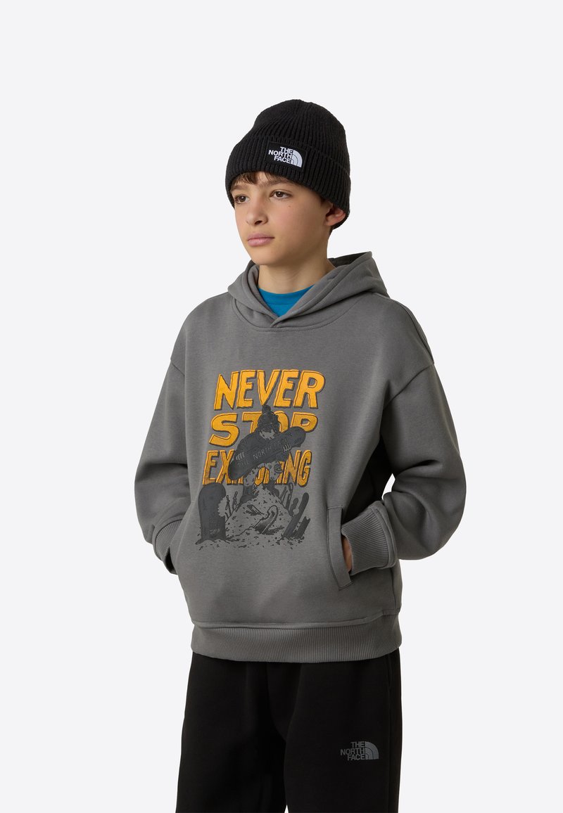 Hall hallipluus, millel on oranži ja musta värvi "NEVER STOP EXPLORING" graafika, kombineeritud musta kootud mütsiga. Modell kannab ka musti pükse.