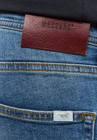 Jeans aus Denim mit einem kastanienbraunen Lederetikett, das mit "MUSTANG" beschriftet ist. Sie verfügen über blaue Nähte, ein weißes Etikett mit einem Logo und eine strukturierte Oberfläche.