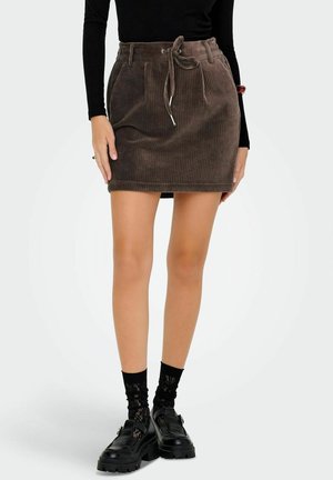 Femme portant une mini-jupe en velours côtelé marron avec cordon, un haut noir à manches longues, des chaussettes noires à motifs et des chaussures noires épaisses, debout sur fond blanc.