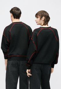 Fekete pulóver piros varrással és raglán ujjakkal, sötét farmerrel párosítva. Laza szabású, bordázott szegélyekkel.