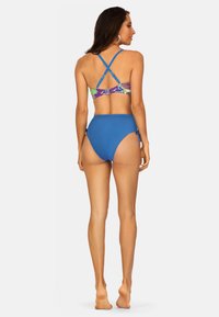 Niebieski strój kąpielowy składający się z wzorzystego biustonosza z krzyżującymi się ramiączkami z tyłu oraz wysokich, gładkich niebieskich majtek. Gładka tekstura materiału.