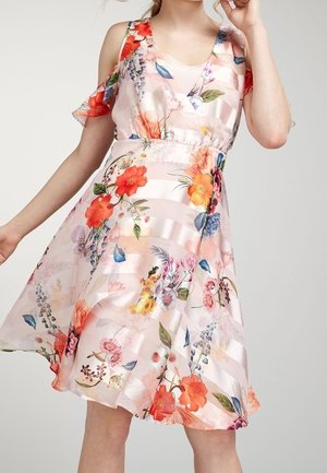 Donna che indossa un abito floreale rosa chiaro con spalle scoperte e gonna fluente, caratterizzato da fiori rossi, arancioni e blu.