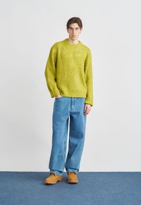 Lys grønn strikkegenser med rund hals, kombinert med løse blå jeans og brune tresko, plassert foran en nøytral bakgrunn.