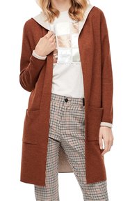 Femme portant un cardigan ouvert couleur rouille, un t-shirt blanc à motif graphique et un pantalon à carreaux taille haute avec fermeture par boutons.