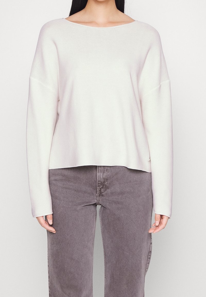 Personne portant un haut blanc ample à manches longues et un jean gris taille haute, debout devant un fond clair uni.