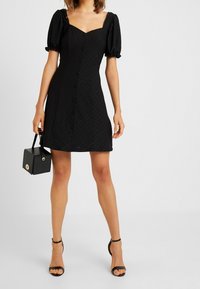 Svart klänning med puffärmar, fyrkantig halsringning och framknäppning. Stylad med svarta sandaler med remmar och en liten svart strukturerad handväska.