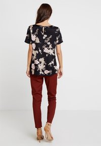 Blouse noire à fleurs avec manches courtes, ornée de fleurs roses et blanches, assortie à un pantalon bordeaux ajusté et des chaussures à talons nus.
