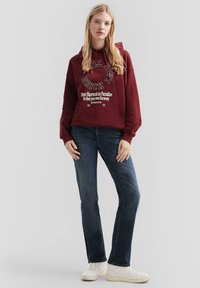 Giovane donna che indossa un hoodie borgogna con testo bianco, jeans blu scuro e sneakers bianche, in piedi contro uno sfondo chiaro e uniforme.