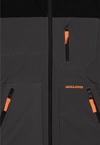 Giacca grigia da uomo con accenti neri, dotata di zip arancioni, tasche sul petto e laterali, e dettaglio con logo nella parte anteriore. Texture liscia.