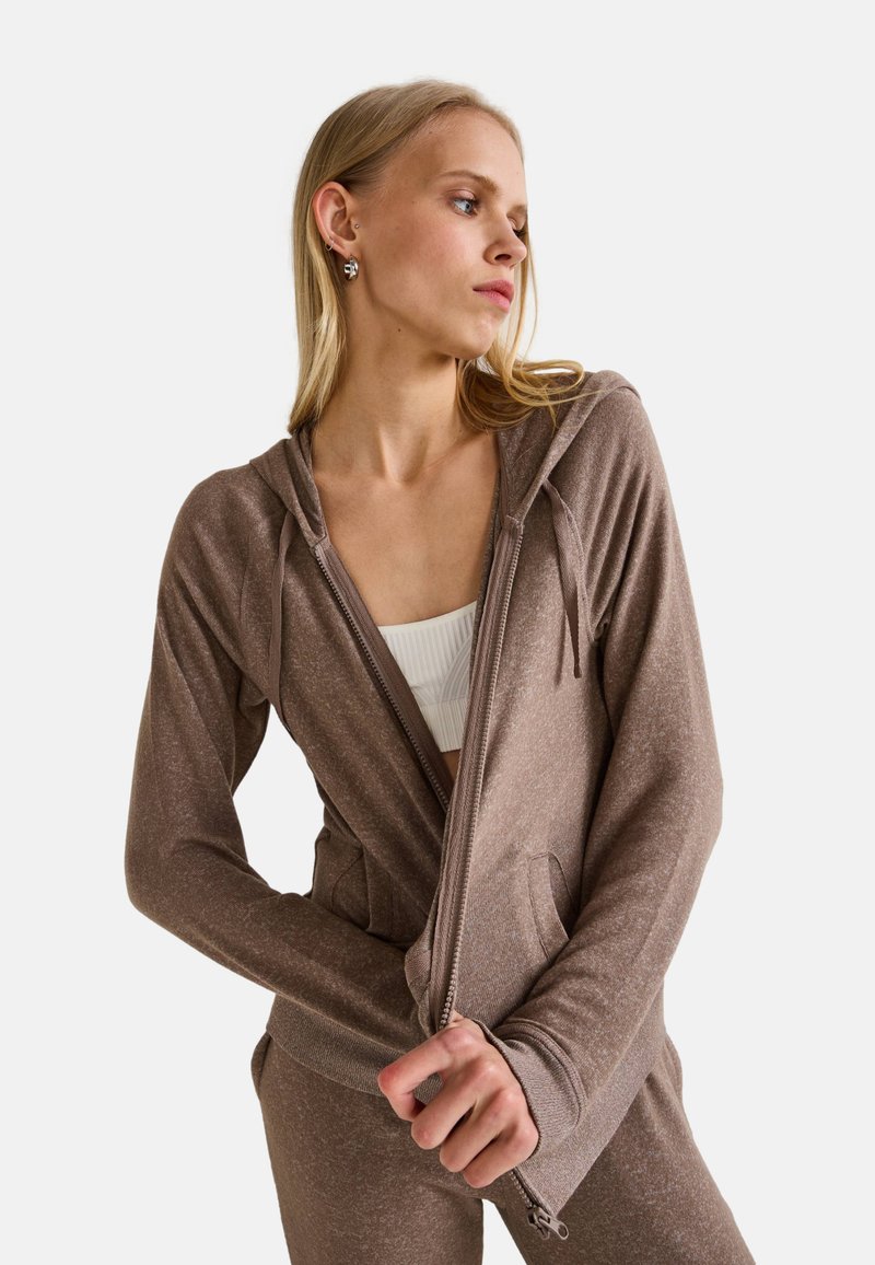 Haut de hoodie zippé marron avec une finition texturée, doté d'une capuche à cordon de serrage, de poches latérales et d'une silhouette ajustée, mettant en valeur un design sportif.