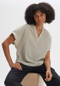 Maglietta grigia chiara a maniche corte con scollatura a V e un motivo a righe texturizzato, indossata con pantaloni neri; posa seduta su un blocco di legno.