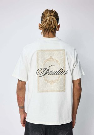 Personne avec des dreadlocks châtain clair attachées en arrière porte un t-shirt blanc avec un motif rectangulaire orné et le mot "Atudios" au dos.