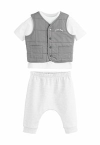 Gilet matelassé gris avec boutons et poches, chemise blanche à manches courtes en dessous, associé à un pantalon de jogging gris clair avec une taille élastique.