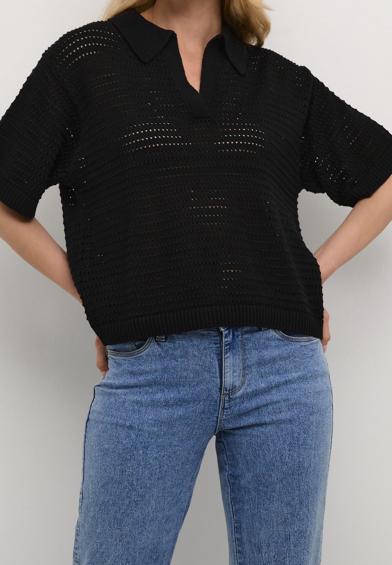 Femme portant un polo crochet noir à manches courtes associé à un jean denim bleu clair taille haute, mains reposant sur les hanches.