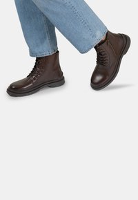 Botas de cuero marrón hasta el tobillo con cordones en la parte delantera, cremallera lateral y suela de goma gruesa. Costuras visibles y una textura suave. Combinadas con denim.