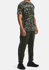 Zelené tričko s maskáčovým vzorem a logem "UNDER ARMOUR", spárované s jednobarevnými zelenými zúženými kalhotami a černými teniskami. Sportovní střih a textilní textura.