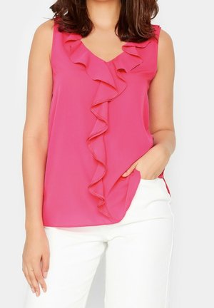 Kvinde iført ærmeløs, skrigpink top med flæser foran kombineret med hvide bukser, hånd i lommen, overkrop og arme synlige.