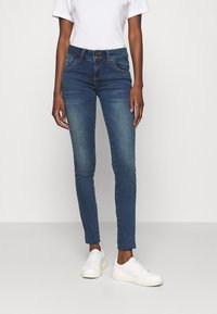 Jeans ajustados en denim azul oscuro con un acabado desgastado, que cuentan con un cierre de cremallera y dos bolsillos frontales. Combinados con zapatillas blancas.