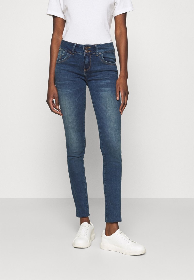 Jeans ajustados en denim azul oscuro con un acabado desgastado, que cuentan con un cierre de cremallera y dos bolsillos frontales. Combinados con zapatillas blancas.