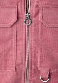 Nahaufnahme eines pinkfarbenen Cordkleidungsstücks mit einem silbernen Reißverschlussanhänger in Form eines Rings und zwei rechteckigen Fronttaschen.