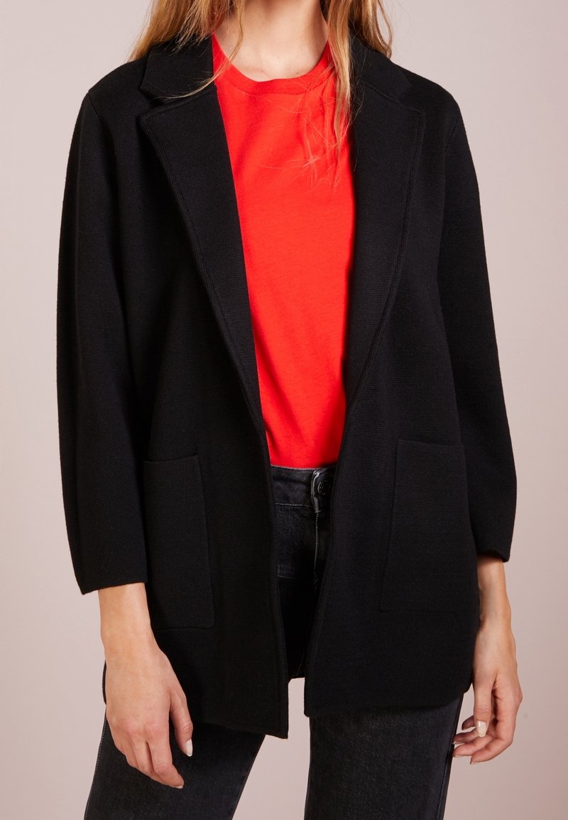 Femme portant un haut rouge vif, une veste noire ouverte avec des poches et un jean foncé, montrée du cou aux cuisses.