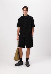 Black short-sleeve polo shirt with textured pattern and matching shorts. Model holds a tan tote bag, paired with dark athletic shoes and socks.