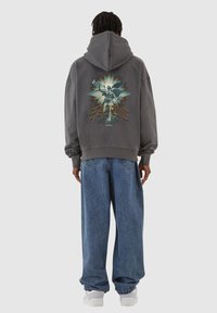 Grå hettegenser med et stort grafisk bilde av en engel og "SAINT"-tekst på baksiden, kombinert med løse, blå denimjeans og hvite joggesko.